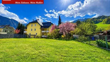 Gelbes Mehrfamilienhaus mit Balkonen und großem Gartenbereich vor einer Bergkulisse unter blauem Himmel.