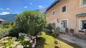 Charmantes Haus mit gepflegtem Garten, blühenden Hortensien und Bergblick unter blauem Himmel.