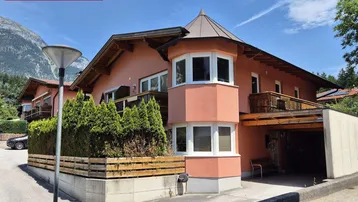 Hausfassade in lachsfarbenem Putz mit Erker und Holzbalkonen vor alpiner Bergkulisse.