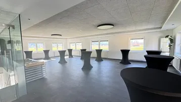 Geräumiger Veranstaltungsraum mit mehreren Stehtischen und großen Fenstern, die viel Tageslicht hereinlassen.