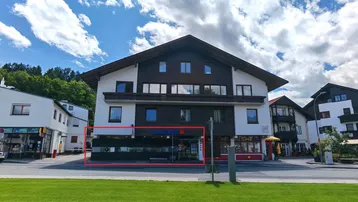Gebäudeansicht von der Straße mit Geschäften im Erdgeschoss und Wohnungen darüber, umgeben von Grünflächen.