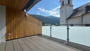 Geräumige Terrasse mit Holzboden, Glasgeländer und malerischem Blick auf Berge und Kirchturm.