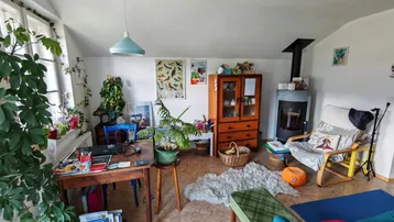 Gemütliches Wohnzimmer mit Holzofen, Schreibtisch und großen Fenstern, die viel Tageslicht hereinlassen.