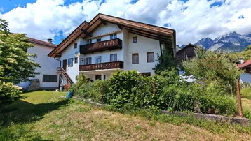 Traditionelles Haus mit Holzbalkonen und Satteldach, umgeben von viel Grün und Bergblick.