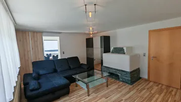Helles Wohnzimmer mit Holzboden, gemütlichem Kachelofen und Zugang zu einem Balkon mit Seeblick.