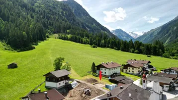 Luftaufnahme eines Dorfes in einem Tal mit Bergen, Wäldern und einem markierten Baugrundstück.