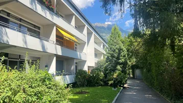 Mehrfamilienhaus mit Balkonen, umgeben von viel Grün und Bergblick unter blauem Himmel.