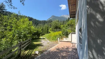 Gepflasterte Terrasse mit kleinem Tisch und Stühlen, umgeben von grüner Natur und Bergblick.