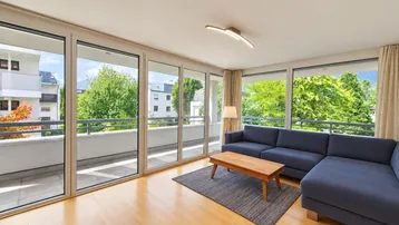 Helles Wohnzimmer mit großen Fensterfronten, Holzboden und Zugang zum Balkon mit Grünblick.