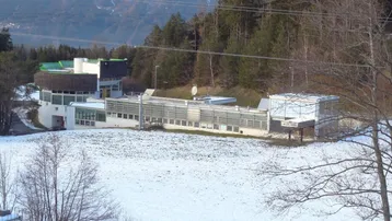 Gebäudekomplex mit runder und langer Struktur, umgeben von Schnee und Bäumen, mit Bergen im Hintergrund.