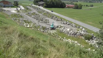 Blick auf ein unbebautes Grundstück mit Schotter und Felsen, umgeben von grünen Wiesen und Bergen, mit einer Landstraße.