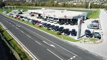 Luftaufnahme eines Autohauses mit großem Parkplatz, umgeben von grünen Feldern und Bergen im Hintergrund.