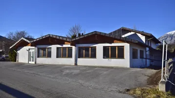 Gebäudeansicht mit weißer Fassade, braunem Dach und Parkplätzen davor, umgeben von Natur und Bergen.