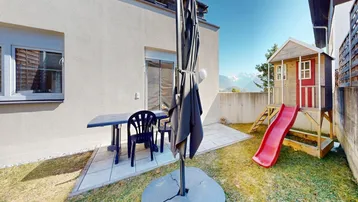 Garten mit gepflasterter Terrasse, Kinderspielhaus und weitem Blick auf die Berge.
