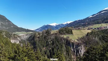 Weitläufige Luftaufnahme einer alpinen Landschaft mit Wäldern, Feldern und schneebedeckten Bergen im Hintergrund.