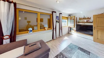 Helles Wohnzimmer mit beigem Sofa, Fernseher und großen Fenstern mit Blick ins Grüne.