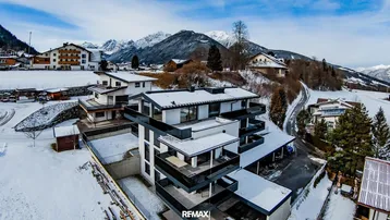 Moderne Wohnanlage in alpiner Landschaft mit schneebedeckten Bergen im Hintergrund und klarem Himmel.