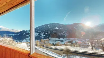 Sonniger Ausblick von einem Holz-Balkon auf eine verschneite Berglandschaft und eine Straße.