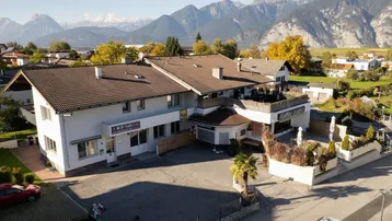 Luftaufnahme eines Gebäudekomplexes mit Bergblick, Parkplätzen und einer Dachterrasse in sonniger Umgebung.