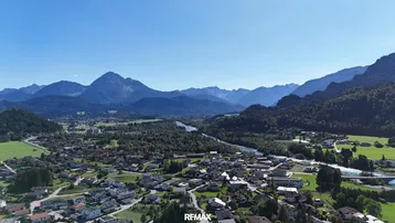 Panoramablick auf eine ländliche Siedlung mit Fluss und majestätischen Bergen im Hintergrund unter blauem Himmel.