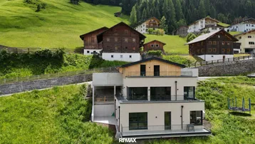Modernes Gebäude in alpiner Landschaft mit traditionellen Häusern im Hintergrund und grünen Hängen.