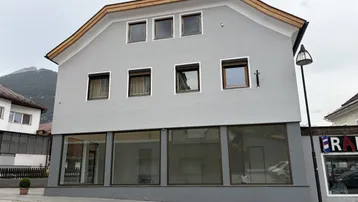 Außenansicht eines Gebäudes mit grauer Fassade, großen Schaufenstern im Erdgeschoss und Wohnfenstern oben.