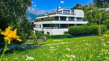 Modernes Mehrfamilienhaus mit Terrassen und Bergblick, umgeben von grüner Wiese und Bäumen.