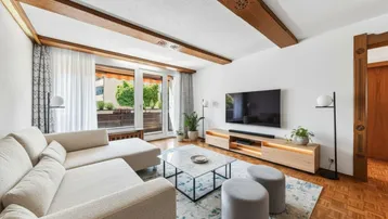 Helles Wohnzimmer mit sichtbaren Holzdeckenbalken, modernem Interieur, Parkettboden und Zugang zu einem Balkon mit Grünblick.
