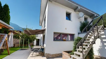 Modernes Haus mit gepflegtem Garten, Terrasse und Außentreppe, umgeben von grüner Landschaft.