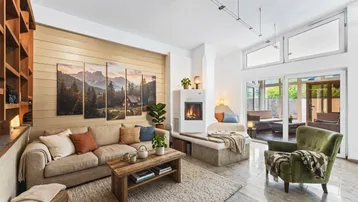 Gemütliches Wohnzimmer mit Kamin, bequemer Couch und Holzwand, hell durch große Fenster zum Außenbereich.