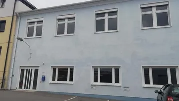 Außenansicht eines hellblauen Gebäudes mit weißen Fenstern, Rollläden und einem kleinen Parkplatz davor.