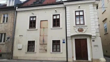 Historisches Gebäude mit klassischer Fassade, braunen Fenstern und rotem Ziegeldach an einer Straße.