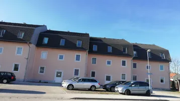 Mehrere Wohngebäude mit hellrosa Fassade und Mansarddächern, umgeben von Parkplätzen.