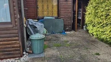 Kleines Holzhaus mit gepflastertem Bereich und angrenzender Hecke unter bewölktem Himmel.