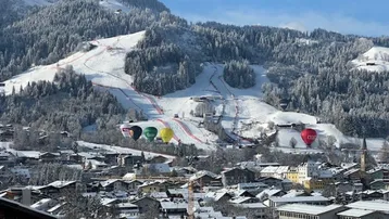 Panoramablick auf eine verschneite Stadt mit Skipisten und Bergen im Hintergrund, Heißluftballons am Himmel.