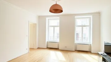 Helles Wohnzimmer mit Parkettboden, hohen Decken und Stuckelementen, durchflutet von natürlichem Licht.