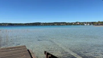 Klarer blauer See mit einem Holzsteg im Vordergrund unter strahlend blauem Himmel.