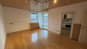 Helles Wohnzimmer mit Parkettboden, Holzdecke und Zugang zu Küche und verglastem Balkon.