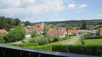Weitläufiger Blick über eine ländliche Siedlung mit Häusern, grünen Hügeln und einem Kirchturm in der Ferne.