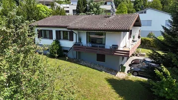 Luftaufnahme eines Einfamilienhauses mit Garten, Balkon und Parkplätzen im Grünen.