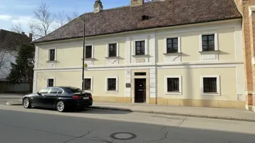 Historisches Gebäude mit beiger Fassade, braunen Fenstern und Satteldach an einer ruhigen Straße.