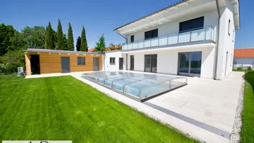 Modernes Haus mit großem Garten, überdachtem Pool und Holz-Nebengebäude unter blauem Himmel.