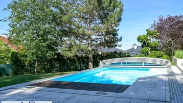 Großer Außenpool mit Abdeckung und Holzdeck, umgeben von gepflegtem Garten und Bäumen.