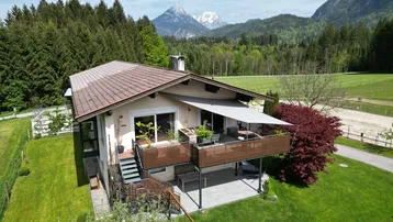 Luftaufnahme eines charmanten Hauses mit großer Terrasse und atemberaubendem Bergblick in grüner Umgebung.
