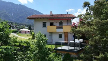 Ansicht des Hauses mit Balkon und Terrasse, umgeben von grüner Natur und Bergen.