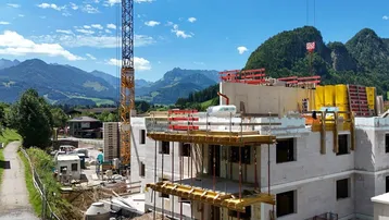 Baustelle eines neuen Wohngebäudes mit Baugerüst und Kran vor einer beeindruckenden Bergkulisse.