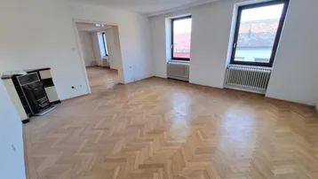 Geräumiges Wohnzimmer mit Fischgrätparkett und traditionellem Kachelofen, hell durch zwei Fenster.