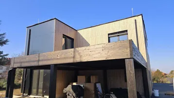 Modernes Haus in Holzbauweise mit Carport und Baustellencharakter unter blauem Himmel.