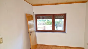Leerer Raum mit Holzboden, Fenster mit Rollladen und Blick ins Grüne.