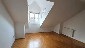 Helles Dachzimmer mit schrägen Wänden, Fenster und Fischgrätparkettboden, ideal als Schlaf- oder Arbeitszimmer.